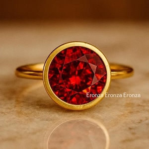 May include: A gold ring featuring a large, faceted, round, red gemstone. The gemstone is set in a gold bezel, and the ring band is also gold. The ring is displayed on a light-colored surface.