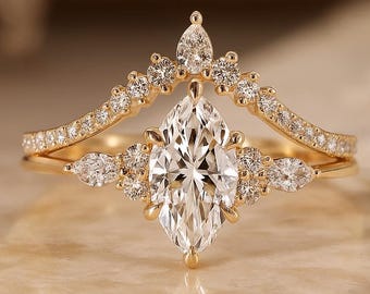 May include: A gold engagement ring set with a marquise-cut diamond centre stone and a diamond-studded band. The ring is set on a marble surface. The band has a curved design with small diamonds.