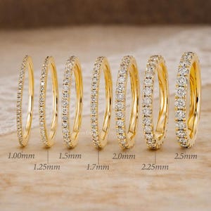 May include: Six gold-colored eternity rings with varying widths, ranging from 1.00mm to 2.5mm, are displayed. Each ring is adorned with small, sparkling gemstones. The rings are arranged on a neutral-toned surface.