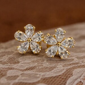 May include: A pair of gold-coloured flower-shaped stud earrings. Each earring features five clear, pear-shaped gemstones arranged to resemble a flower. The earrings are set against a soft, lace-like background.
