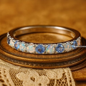 May include: A silver ring featuring alternating white opal and blue gemstones. The ring is displayed on a wooden surface with a lace doily. The gemstones are round and evenly spaced around the band.