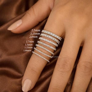 May include: A collection of diamond eternity rings of varying widths, from 1.00mm to 2.5mm, displayed on a finger. The rings are set with small, sparkling stones, creating a continuous band of brilliance. The rings are shown against a neutral background.