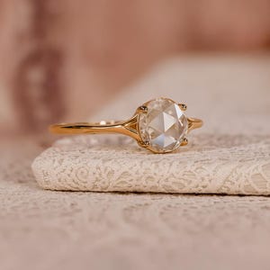 May include: A gold ring featuring a large, oval-cut, clear gemstone. The ring has a slender band with a split design near the stone. The gemstone is faceted, reflecting light and creating a sparkling effect. The ring is displayed on a white, textured surface.