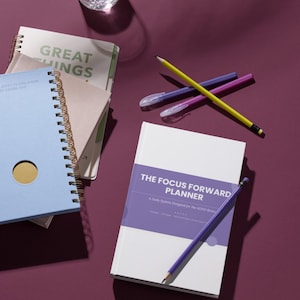 May include: A white planner titled The Focus Forward Planner with a purple band, alongside a light blue spiral notebook, a notebook with the text Great Things, a yellow pencil, and two purple pens. The items are arranged on a dark purple surface.