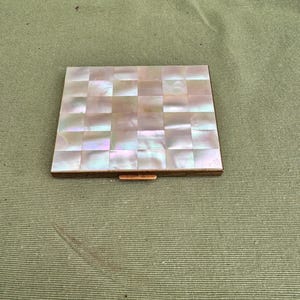 Puede incluir: Un estuche compacto cuadrado y vintage con un diseño a cuadros de nácar. Los cuadrados iridiscentes están engastados en un marco de metal dorado. El estuche compacto está cerrado, con un pequeño cierre visible.