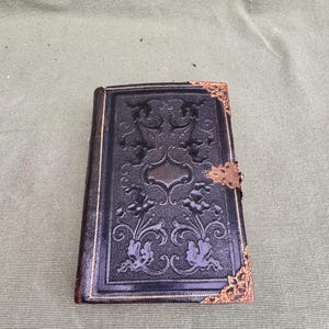 May include: A small, antique black book with ornate floral embossed detailing. The cover features a decorative brass clasp and corner accents. The book is closed and appears to be made of leather or a similar material.