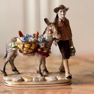 Puede incluir: Figura de cerámica que representa a un hombre con un burro. El burro gris tiene adornos coloridos y lleva cestas. El hombre lleva sombrero y camisa marrones y sostiene un cántaro. Las cestas están llenas de objetos.