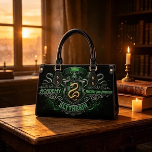 May include: Black handbag with a green and gold Slytherin crest design. The bag features a top handle and the words "Academy", "Slytherin", "Mystery and Ambition". The bag is sitting on a wooden table.