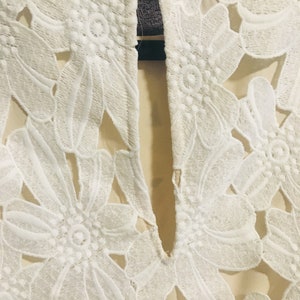 May include: White lace fabric with a floral pattern. The fabric has a sheer look and is cut out in the shape of flowers.
