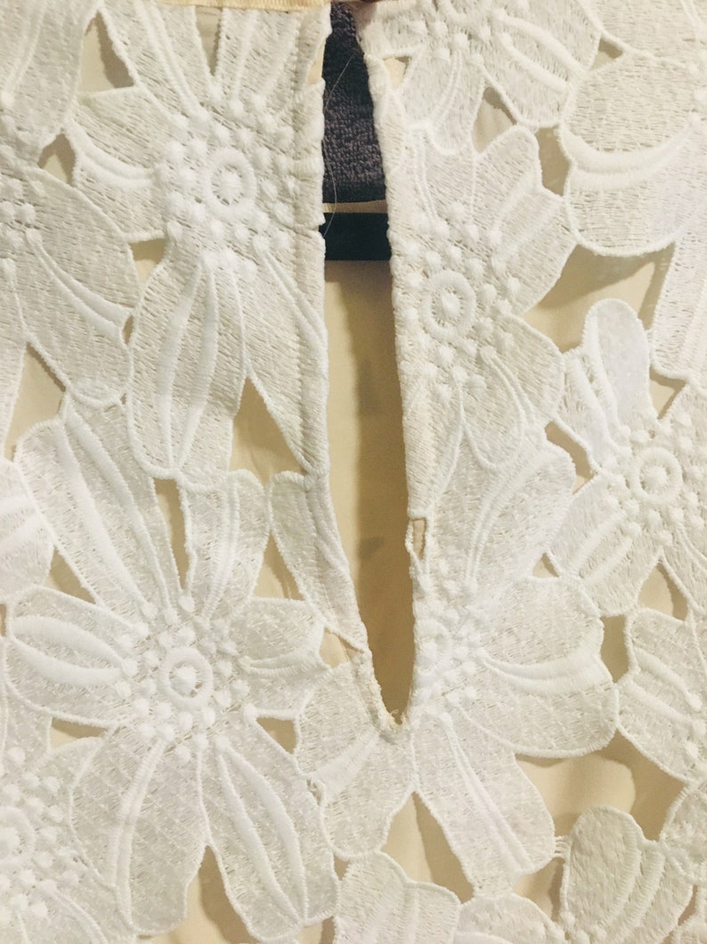May include: White lace fabric with a floral pattern. The fabric has a sheer look and is cut out in the shape of flowers.