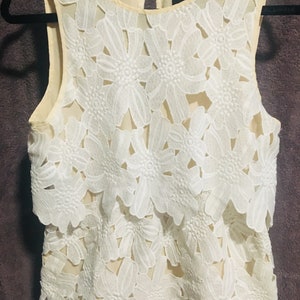 May include: A white sleeveless top with a layered floral lace design. The top is made of a sheer fabric and has a scalloped hem.