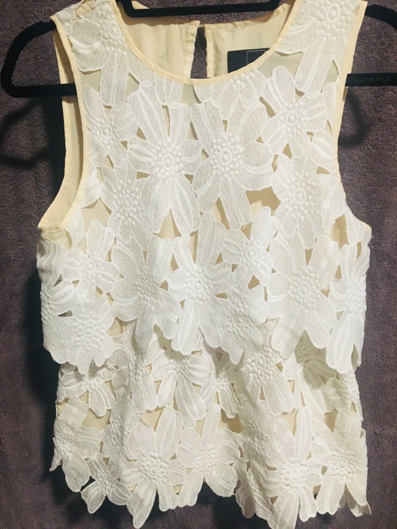May include: A white sleeveless top with a layered floral lace design. The top is made of a sheer fabric and has a scalloped hem.