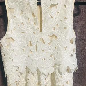May include: A white sleeveless top with a delicate floral lace pattern. The top has a round neckline and a keyhole opening at the back.