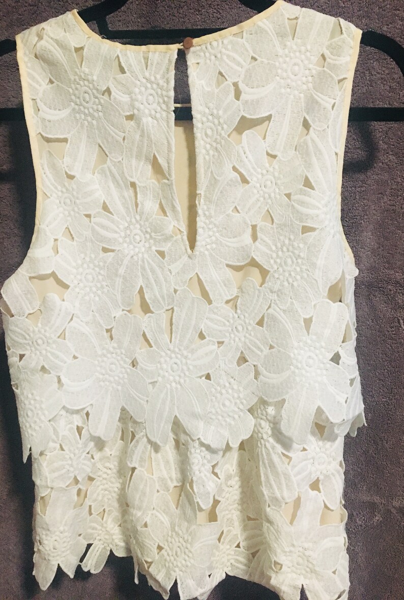 May include: A white sleeveless top with a delicate floral lace pattern. The top has a round neckline and a keyhole opening at the back.