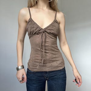 May include: A brown camisole top with spaghetti straps and a V-neckline. The top features a tie detail at the bust and a delicate eyelet pattern throughout the fabric. The garment is paired with blue denim jeans.