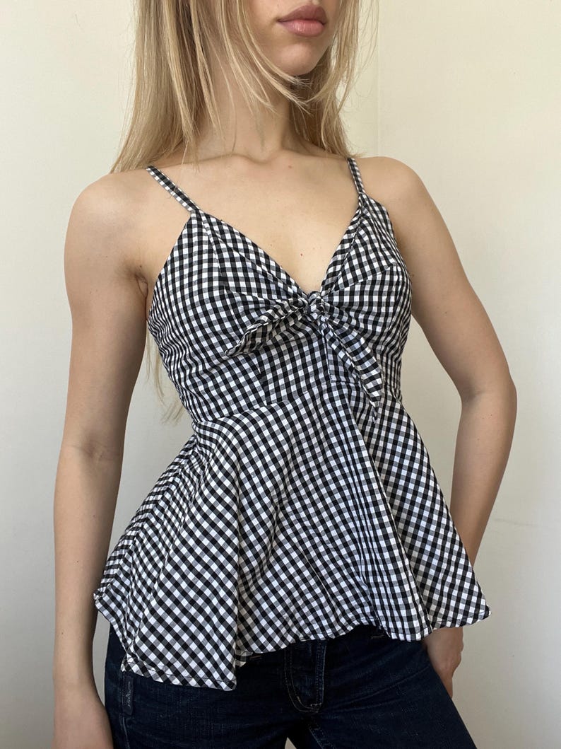 May include: A black and white gingham print camisole top with spaghetti straps and a bow detail at the bust. The top has a peplum style, flaring out from the waist. The fabric is a classic checkered pattern.