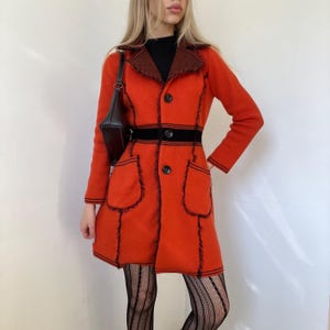 Puede incluir: Un abrigo naranja vintage con ribetes negros y un cinturón negro. El abrigo presenta dos bolsillos delanteros y un cuello marrón en contraste. El abrigo de manga larga tiene dos botones negros y un bolso negro.