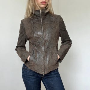 Può includere: Una giacca marrone con un motivo testurizzato a pelle di serpente sui pannelli frontali e maniche marroni a tinta unita. Il capo presenta un colletto alto, una chiusura a zip integrale in metallo argentato e due tasche laterali con zip, indossata sopra jeans blu.