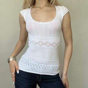 May include: A white knit top featuring a scoop neckline and cap sleeves. The garment displays a variety of textured patterns, including vertical ribbing, horizontal bands, and a central row of diamond-shaped openwork detailing across the midsection.