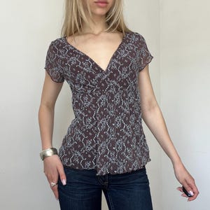 May include: A brown V-neck top with a delicate white floral pattern. The short-sleeved blouse has a fitted bodice and a subtle lace trim along the neckline. The fabric appears lightweight and semi-sheer, suitable for warmer weather.