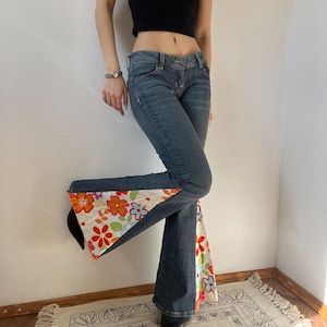 May include: Blue denim jeans with a flared leg design. The jeans feature a floral pattern in shades of orange, red, pink, and green on a white background at the flared hem. The jeans have a classic five-pocket design.