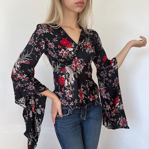 May include: A black long-sleeve top featuring a vibrant red and white floral pattern with a v-neckline and wide, flared bell sleeves. The garment is paired with blue denim jeans, showcasing a fitted silhouette and a detailed botanical print.