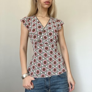 May include: A sleeveless wrap top with a white background and a repeating pattern of brown, pink, and black circles. The top has a V-neckline and a fitted silhouette. The edges are trimmed with a dark brown color.
