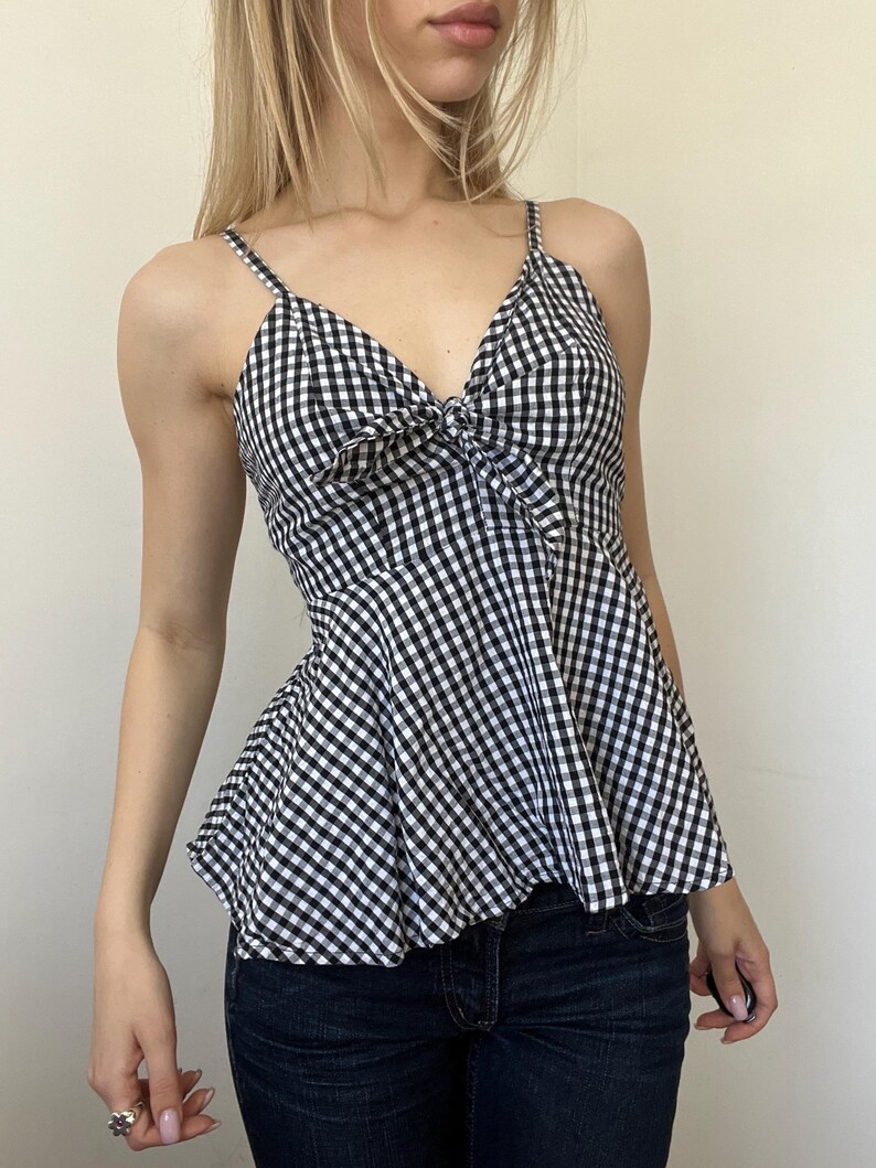 May include: A black and white gingham print camisole top with spaghetti straps and a bow detail at the bust. The top has a peplum-style hem and is paired with dark blue denim jeans.