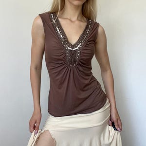 May include: A sleeveless brown top featuring a deep V-neckline adorned with intricate silver and dark beadwork and sequins. The fabric is gathered at the center of the chest, creating soft vertical pleats that extend downward across the bodice.