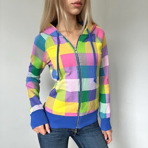 May include: A zip-up hoodie featuring a vibrant multi-colored plaid pattern in shades of yellow, pink, green, purple, and blue. The garment includes a hood with blue drawstrings, a full-length metal zipper, and solid blue ribbed cuffs and hem.