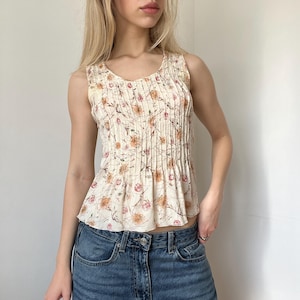 May include: Cream-colored sleeveless top with a floral pattern in shades of orange and pink. The top features vertical pleats down the front and a rounded neckline. The fabric appears lightweight and suitable for warm weather.