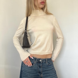 Puede incluir: Jersey de manga larga color crema con cuello alto y dobladillo festoneado. El jersey se combina con vaqueros azules. También se ve un pequeño bolso negro.