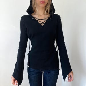 May include: A black long-sleeve hooded top featuring a V-neckline with a lace-up cord detail, flared sleeves, and side lacing accents with silver-toned metal hardware, paired with dark blue denim jeans.