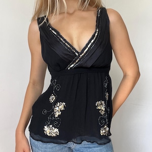May include: A black sleeveless top featuring a deep V-neckline with silver sequin trim. The bodice has a gathered empire waist and is decorated with floral-inspired silver sequin embroidery on the lower front panels. The fabric has a textured finish.