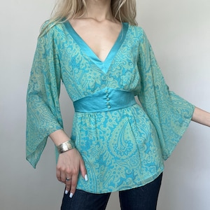 May include: A turquoise paisley print blouse with wide sleeves and a V-neck. The blouse features a turquoise satin band at the waist with decorative buttons. The blouse is made of a lightweight, flowing fabric.