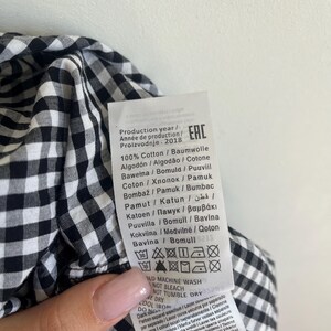 May include: Close-up of a black and white gingham fabric with a care label attached. The label includes washing instructions and fabric composition details. The fabric is 100% cotton and the production year is 2018.