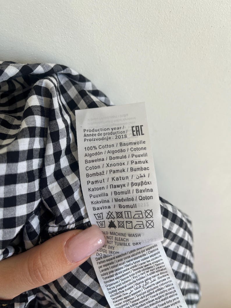 May include: Close-up of a black and white gingham fabric with a care label attached. The label includes washing instructions and fabric composition details. The fabric is 100% cotton and the production year is 2018.