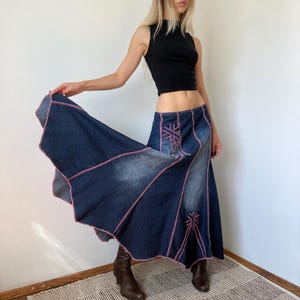 May include: A long, flared denim skirt featuring a dark blue wash with faded areas, accented by prominent pink contrast stitching along the panel seams and decorative geometric embroidery patterns in pink thread on the front panels.