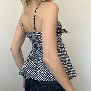 May include: A black and white gingham print camisole top with adjustable spaghetti straps and a peplum hem. The top features a bow detail at the side and a smocked back panel for a comfortable fit. The top is paired with dark wash denim jeans.