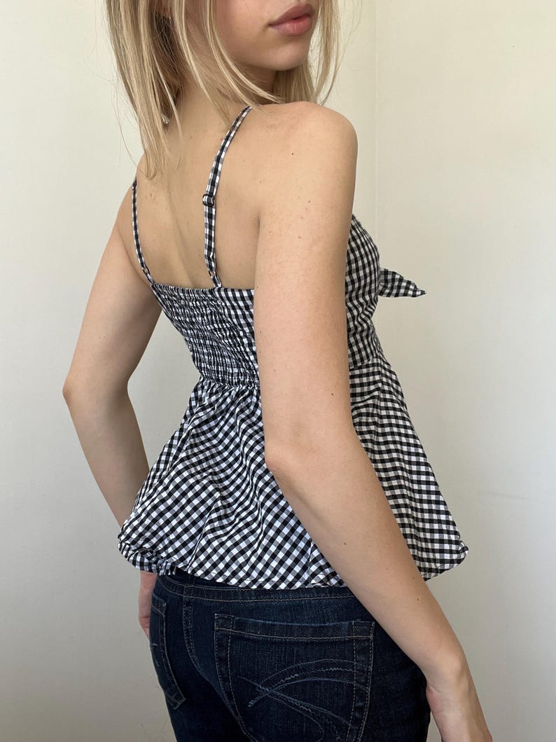 May include: A black and white gingham print camisole top with adjustable spaghetti straps and a peplum hem. The top features a bow detail at the side and a smocked back panel for a comfortable fit. The top is paired with dark wash denim jeans.