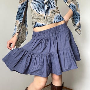 Könnte beinhalten: Ein blauer, gestufter Minirock mit elastischem Bund und geraffter, ausgestellter Silhouette. Der Rock wird mit einem langärmeligen Oberteil kombiniert, das ein blau-beiges Tiermuster, ausgestellte Ärmel und ein Paisley-Design aufweist.