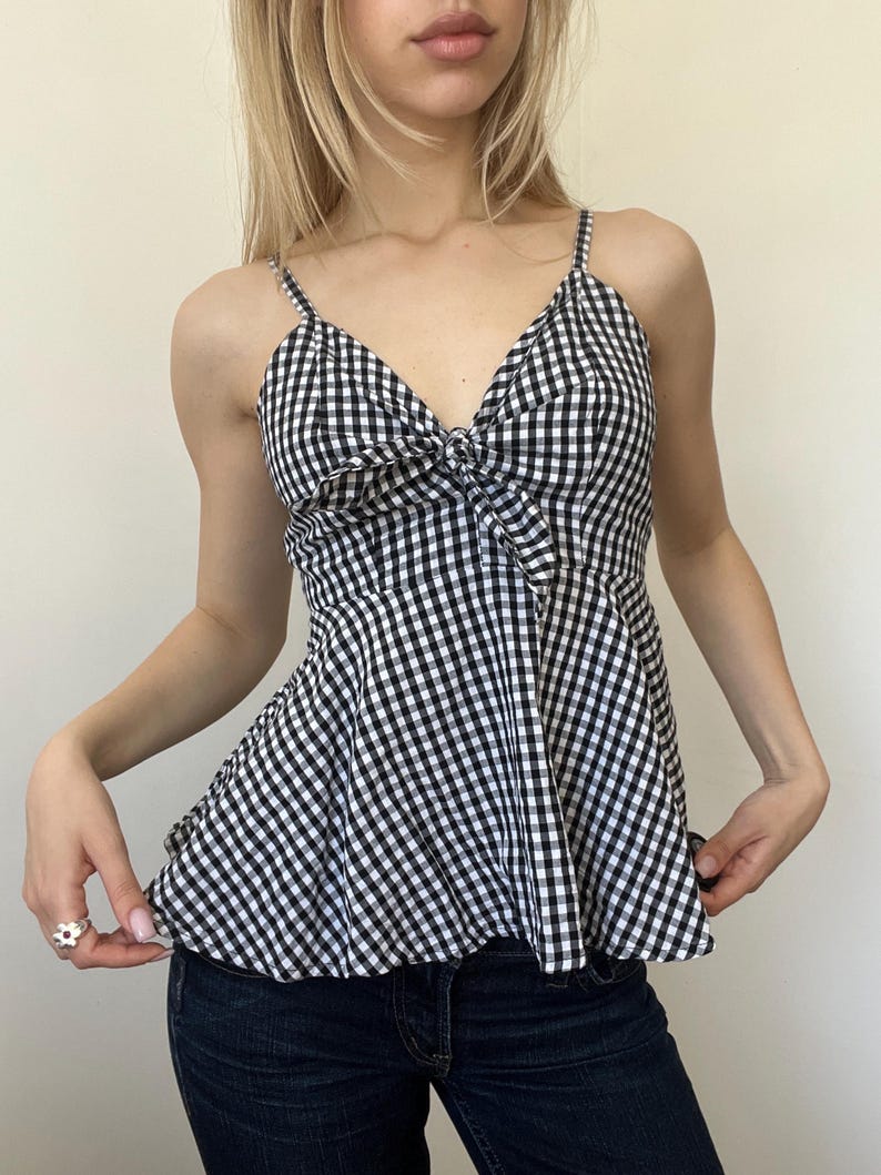May include: A black and white gingham print top with spaghetti straps and a bow detail at the bust. The top has a peplum style and is paired with dark wash denim jeans. The top is a classic, feminine style.