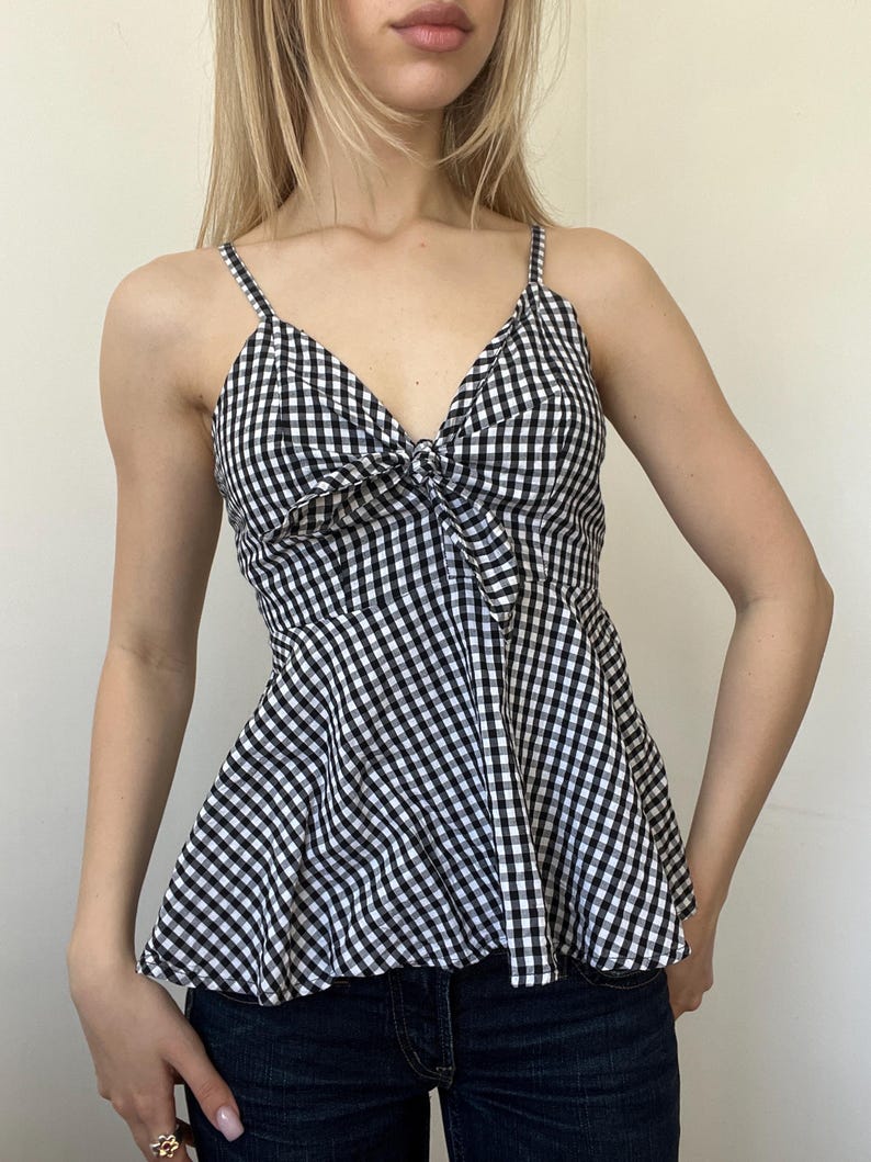 May include: A black and white gingham print camisole top with spaghetti straps and a bow detail at the bust. The top has a peplum-style hem and is paired with dark wash denim jeans.
