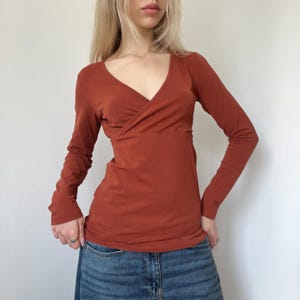 May include: Rust-colored long-sleeve top with a wrap-style front and a V-neckline. The top is made of a soft, form-fitting material. The sleeves are long and fitted, and the overall design is simple and elegant.