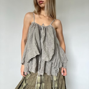 Peut inclure: Un haut caraco sans manches de couleur taupe présentant un design ample, superposé et à volants. Le vêtement possède de fines bretelles et une encolure froncée avec un petit détail de lien central. Le tissu a un aspect texturé et froissé.