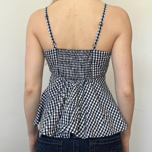 May include: A black and white gingham print top with adjustable spaghetti straps. The top has a fitted, smocked bodice and a peplum-style hem. The straps and bodice are black and white checkered. The top is paired with dark wash denim jeans.