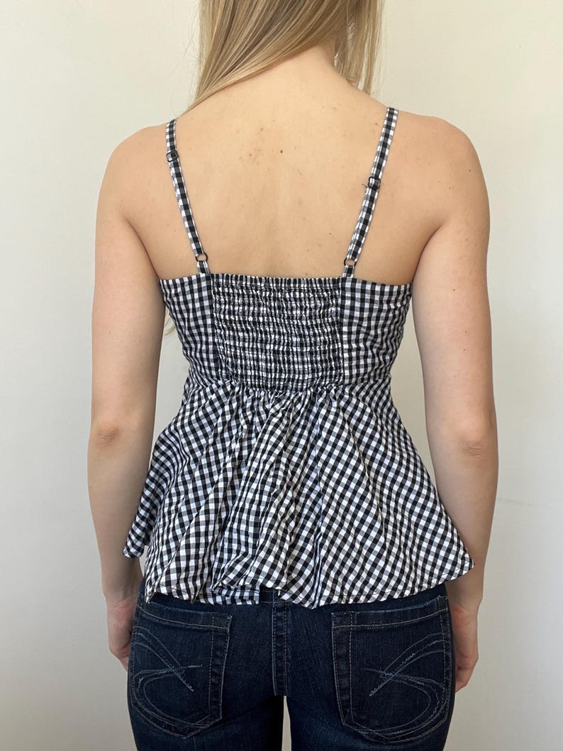 May include: A black and white gingham print top with adjustable spaghetti straps. The top has a fitted, smocked bodice and a peplum-style hem. The straps and bodice are black and white checkered. The top is paired with dark wash denim jeans.