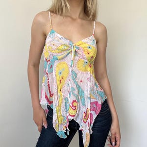 May include: A sleeveless camisole top featuring a white base with a colorful paisley print in shades of yellow, turquoise, and magenta. The garment has thin spaghetti straps, a gathered center front with a tie detail, and an asymmetrical hemline.