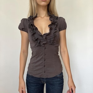 Puede incluir: Una blusa de color gris oscuro con mangas cortas y un escote en V profundo adornado con un volante prominente. La prenda entallada incluye una fila vertical central de pequeños botones a juego que van desde la cintura hasta el cuello.