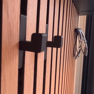Könnte beinhalten: Holzverkleidung mit vertikalen Holzlatten und schwarzen Akzenten. Zwei schwarze, quadratische Haken sind an der Verkleidung angebracht. Eine Brille mit silbernem Rahmen hängt an der angrenzenden Wand.